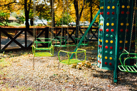 Green swings hang motionless in a quiet park surrounded by trees showcasing autumn foliage inviting...