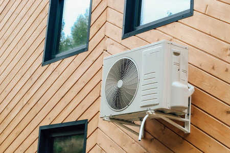 A sleek air conditioning unit is mounted on the wooden facade of a modern house. Sunlight highlights...
