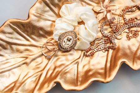 Delicate rings a satin scrunchie and sparkling necklaces rest beautifully on a golden leaf tray. This stylish arrangement showcases various accessories in an elegant setting.の写真素材