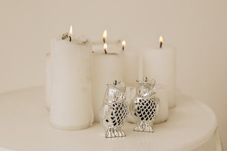 Two shiny silver owl figurines rest near softly glowing white candles on a white table. The scene creates a peaceful atmosphere ideal for relaxation or meditation enhancing the calm environment.の写真素材