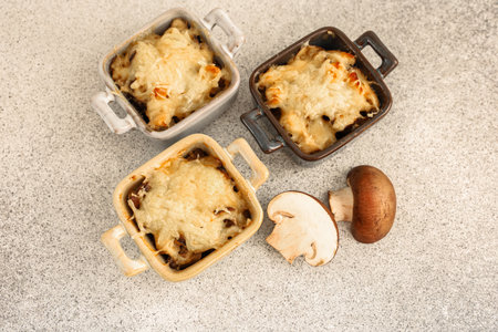 Three mini casserole dishes hold baked mushroom Julienne meals topped with cheese. Fresh mushrooms are placed next to the dishes showcasing a delicious home cooked meal ready to enjoy.の写真素材