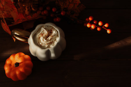 A warm drink topped with whipped cream and spices sits in a pumpkin shaped mug. Autumn leaves and decorative pumpkins add seasonal charm to the rustic wooden table.の写真素材