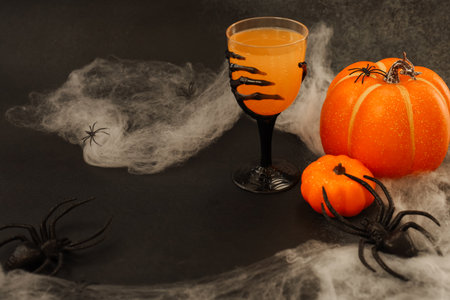 Colorful Halloween scene features a black goblet filled with orange drink beside bright pumpkins and fake spiders. Spooky cobwebs create an eerie atmosphere perfect for a Halloween gathering.の写真素材