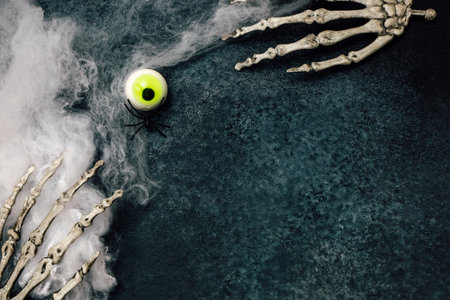 A spooky Halloween decoration displays two skeleton hands arranged on a dark surface with a bright green eyeball in the center. Cobwebs add to the eerie atmosphere of the scene.の写真素材