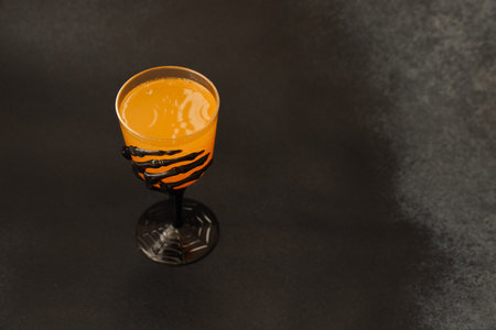 A bright orange beverage is served in a black glass decorated with a spider web and skeleton hand. The drink adds a festive touch to a dark table setting ideal for Halloween celebrations.の写真素材