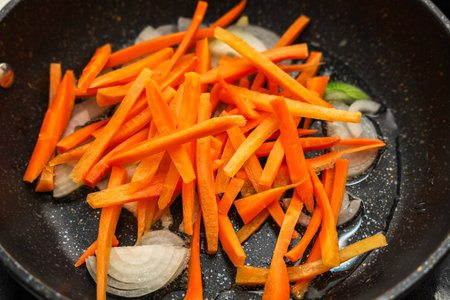 Carrots are cut into thin strips and mixed with onion slices cooking in a skillet on medium heat. The scene shows a vibrant display of color and fresh vegetables typical of home cooking.の写真素材