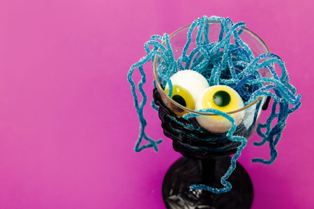 A spooky dessert featuring two candy eyeballs resting in a glass filled with bright blue sugary strands. The setting creates a festive Halloween vibe with a vibrant purple background.の写真素材