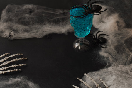 A dark table is set for Halloween with a blue drink in a fancy glass surrounded by plastic spiders and skeleton hands. Cobwebs add to the eerie atmosphere creating a festive spooky vibe.の写真素材