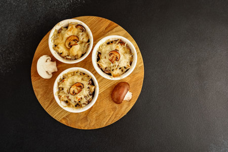 Three ramekins filled with baked mushroom dishes Julienne are arranged on a round wooden board. The meal is presented simply with fresh mushrooms beside the dishes highlighting the flavors.の写真素材