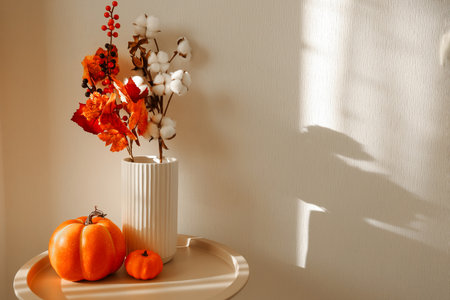 A lovely display of autumn decor featuring vibrant orange pumpkins and white cotton flowers in a textured vase. The soft light casts gentle shadows enhancing the cozy atmosphere in the room.の写真素材