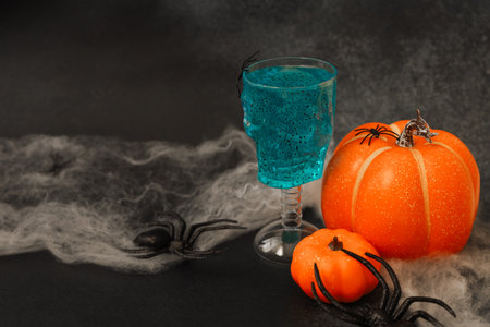 A bright blue drink sits in a glass shaped like a skull surrounded by two pumpkins one orange and one small. Black spiders creep along a web like backdrop creating a Halloween theme.の写真素材