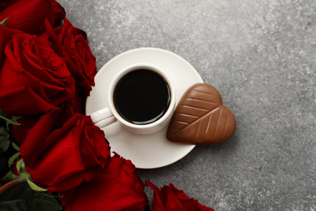 A cup of black coffee sits on a white saucer beside a heart shaped chocolate cookie surrounded by vibrant red roses on a gray stone table creating a romantic atmosphere for any occasion.の写真素材