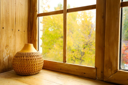 Sunlight fills a cozy room illuminating a beautifully crafted woven vase resting on a wooden windowsill. Outside vibrant autumn leaves provide a colorful backdrop enhancing the serene atmosphere.の写真素材