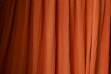 Soft orange fabric hangs gracefully in folds capturing light and shadow. Its texture adds depth to the scene suggesting a cozy space. The warm color evokes feelings of comfort and warmth.の写真素材