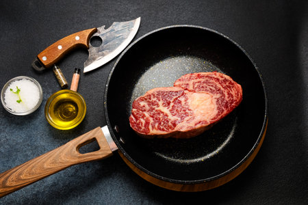 A thick marbled steak is placed in a frying pan ready to be cooked. Surrounding the pan are fresh herbs oil and a knife. The scene is set on a dark surface evoking a culinary atmosphere.の写真素材