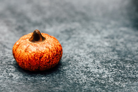A small orange pumpkin is placed on a dark textured surface. The pumpkin has a smooth skin with notable ridges and a dark brown stem. Its vibrant color offers a warm contrast against the background.の写真素材