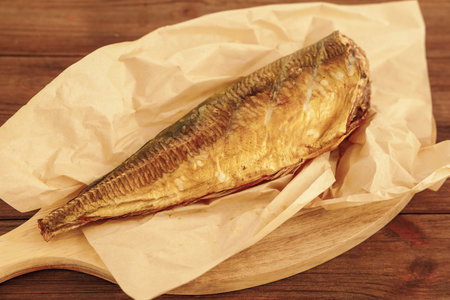 whole fish smoked mackerel is presented on a wooden board resting on crumpled brown paper. The fish is golden brown from smoking and looks delicious ready to be served for a meal.の写真素材