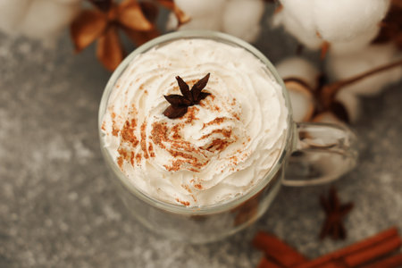 A warm beverage sits in a clear mug featuring rich whipped cream topped with a sprinkle of cinnamon and anise star. Cotton and spices add a cozy touch to the scene perfect for chilly days.の写真素材