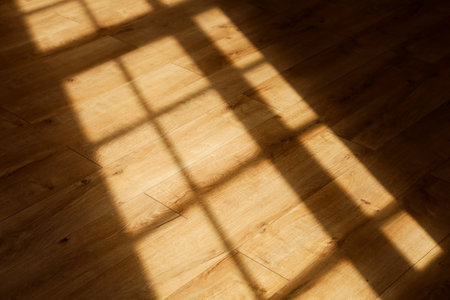 Bright sunlight streams through a window casting geometric shadows on a smooth wooden floor in the afternoon. The warm tones of the wood contrast with the dark shapes created by the light.の写真素材