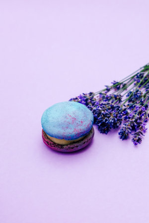 A vibrant macaron sits on a soft pink surface next to fresh lavender flowers. The macaron features bright colors adding a playful touch to the serene arrangement.の写真素材