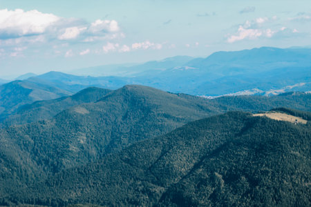 Rolling green hills and valleys stretch across the landscape with distant mountains fading into the horizon. Soft clouds dot the bright blue sky creating a peaceful scene in nature.の写真素材