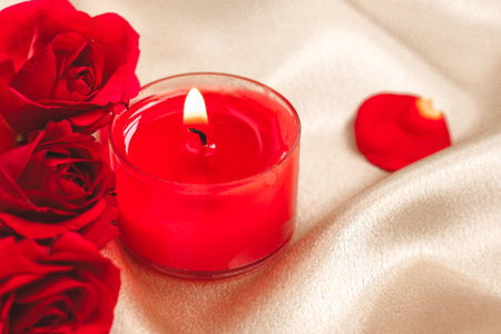 A single red candle flickers beside vibrant red roses arranged gracefully. Satin fabric drapes softly in the background enhancing the romantic atmosphere with scattered rose petals.の写真素材