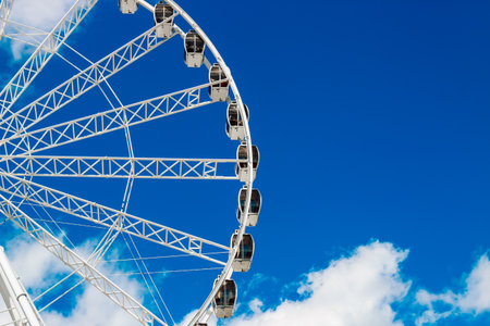 A large ferris wheel stands against a clear blue sky showcasing its white cabins. The sunny day highlights the structure inviting visitors to enjoy a scenic ride above the park.の写真素材