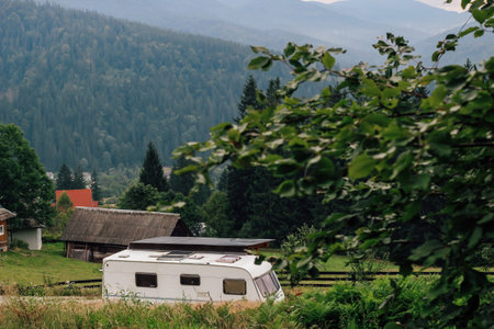 A camper van sits peacefully in a lush green meadow framed by trees. Nearby rustic houses contribute to the serene atmosphere with mountains rising in the background under a clear sky.の写真素材