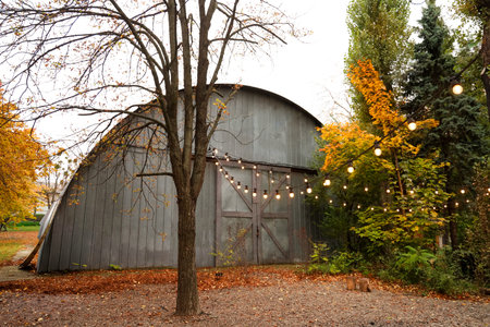 A large curved barn sits in a peaceful outdoor area surrounded by vibrant fall leaves. String lights...