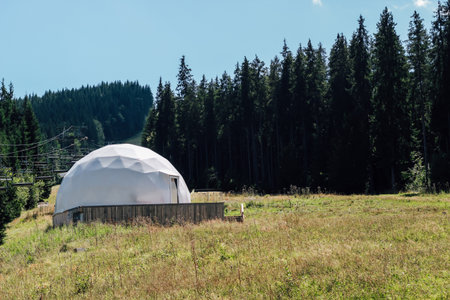 A distinctive dome shaped building stands alone in a lush green field surrounded by tall trees. The bright blue sky and warm sunlight create a tranquil atmosphere in the natural landscape.の写真素材