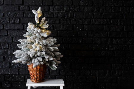 A small Christmas tree dusted with white and adorned with warm lights stands in a woven basket. The dark brick wall behind it adds a cozy festive atmosphere perfect for holiday celebrations.の写真素材