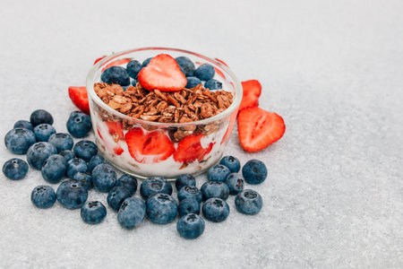 A clear bowl filled with yogurt topped with granola fresh strawberries and blueberries sits on a...