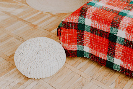 A warm and inviting living area features a red and green plaid blanket draped over a cushioned seat. Beside it sits a round white cushion on a light wooden floor creating a homey atmosphere.の写真素材