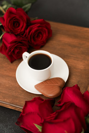 Rich black coffee served in a white cup accompanies a chocolate treat on a wooden board surrounded by bright red roses. This setup creates a warm and inviting atmosphere for enjoying a sweet moment.の写真素材