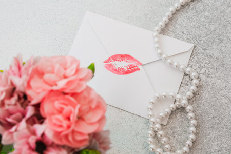 A white envelope with a red lipstick kiss mark sits on a smooth grey surface. Nearby a bouquet of pink flowers and a string of white pearls add elegance to the scene. The setting looks romantic.の写真素材
