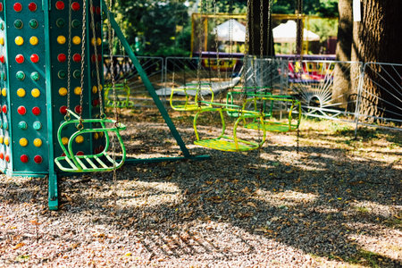 Swings hang still in an amusement park surrounded by tall trees and soft gravel ground. The colorful structures hint at a lively atmosphere now peaceful in the fading afternoon light.の写真素材