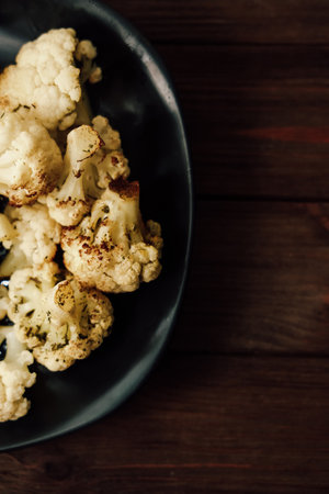 Freshly roasted cauliflower pieces are arranged neatly on a black plate. The dish features a golden brown color and is placed on a rustic wooden table creating a warm inviting atmosphere.の写真素材