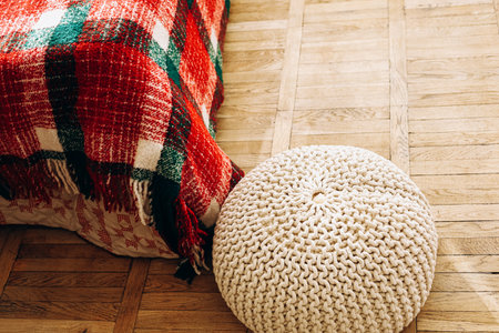 A cozy room features a red and green checked blanket draped over a bed and a round knitted pouf resting on a warm wooden floor. The scene suggests comfort and relaxation in a stylish setting.の写真素材