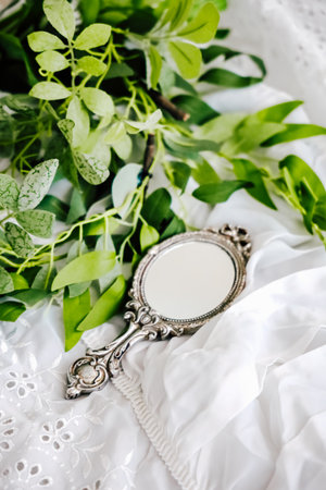 A vintage mirror rests among lush green leaves on a soft white fabric.の写真素材