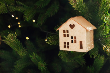 Small wooden house ornament with a heart shaped cutout hangs on a Christmas tree surrounded by green pine branches and soft glowing lights creating a festive atmosphere during the holiday season.の写真素材