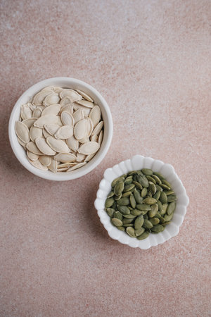 Two small bowls contain pumpkin seeds. One bowl holds roasted green seeds while the other has raw white seeds. The scene is set on a soft light surface ideal for cooking or snacking.の写真素材