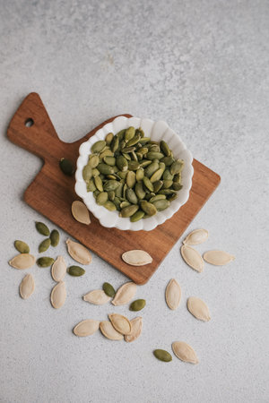 A small white bowl filled with green pumpkin seeds sits on a wooden cutting board. Some seeds are scattered around on a light gray surface.の写真素材