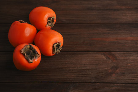 Four ripe persimmons sit on a rustic wooden surface. Their bright orange skin contrasts beautifully with the dark wood creating a warm and inviting scene.の写真素材