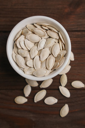 A white bowl filled with pumpkin seeds sits on a wooden surface. Some seeds are scattered around highlighting their flat oval shape and smooth texture under natural lighting.の写真素材