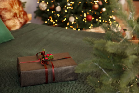 A wrapped gift with decorative bow rests on a green bedspread surrounded by a vibrant Christmas tree...