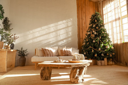 A warm and inviting living room features a beautifully decorated Christmas tree. Sunlight streams through large windows illuminating comfortable seating and rustic wooden furniture.の写真素材