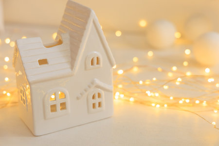 A small white ceramic house is set against a softly glowing backdrop of warm fairy lights. The scene creates a cozy atmosphere suitable for winter decorations and festive gatherings.の写真素材