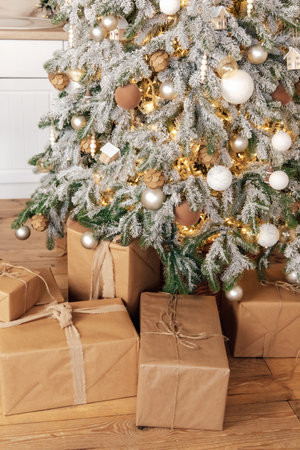 A charming Christmas tree stands tall adorned with golden and white ornaments. Below wrapped gifts in brown paper sit adding to the festive atmosphere of a warm living room.の写真素材