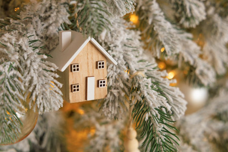 A charming wooden house ornament is suspended from a frosted pine branch. The festive background includes soft lights creating a warm holiday atmosphere. This scene captures the joy of the season.の写真素材