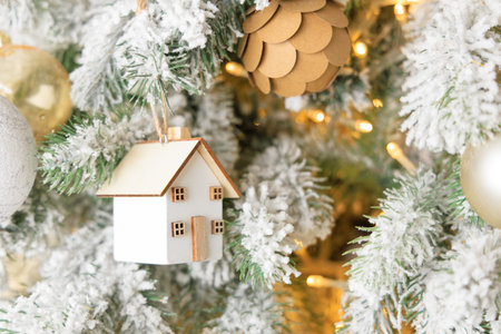A charming white house ornament is suspended from a snow covered Christmas tree. The tree is adorned with golden accents and soft glowing lights creating a warm holiday atmosphere.の写真素材